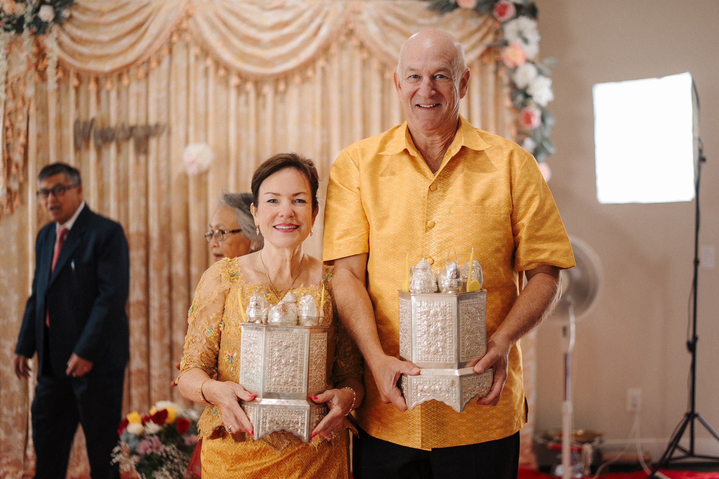 People in matching gold outfits holding gift boxes at an elegant party with draped curtains and decorations.