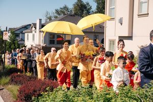 Buddhist monks in orange and yellow robes walk in a processional line under bright yellow umbrellas on a sunny day.
