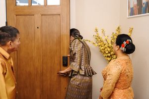 Three people gather near a wooden door wearing formal vintage-inspired garments in brown and orange tones.