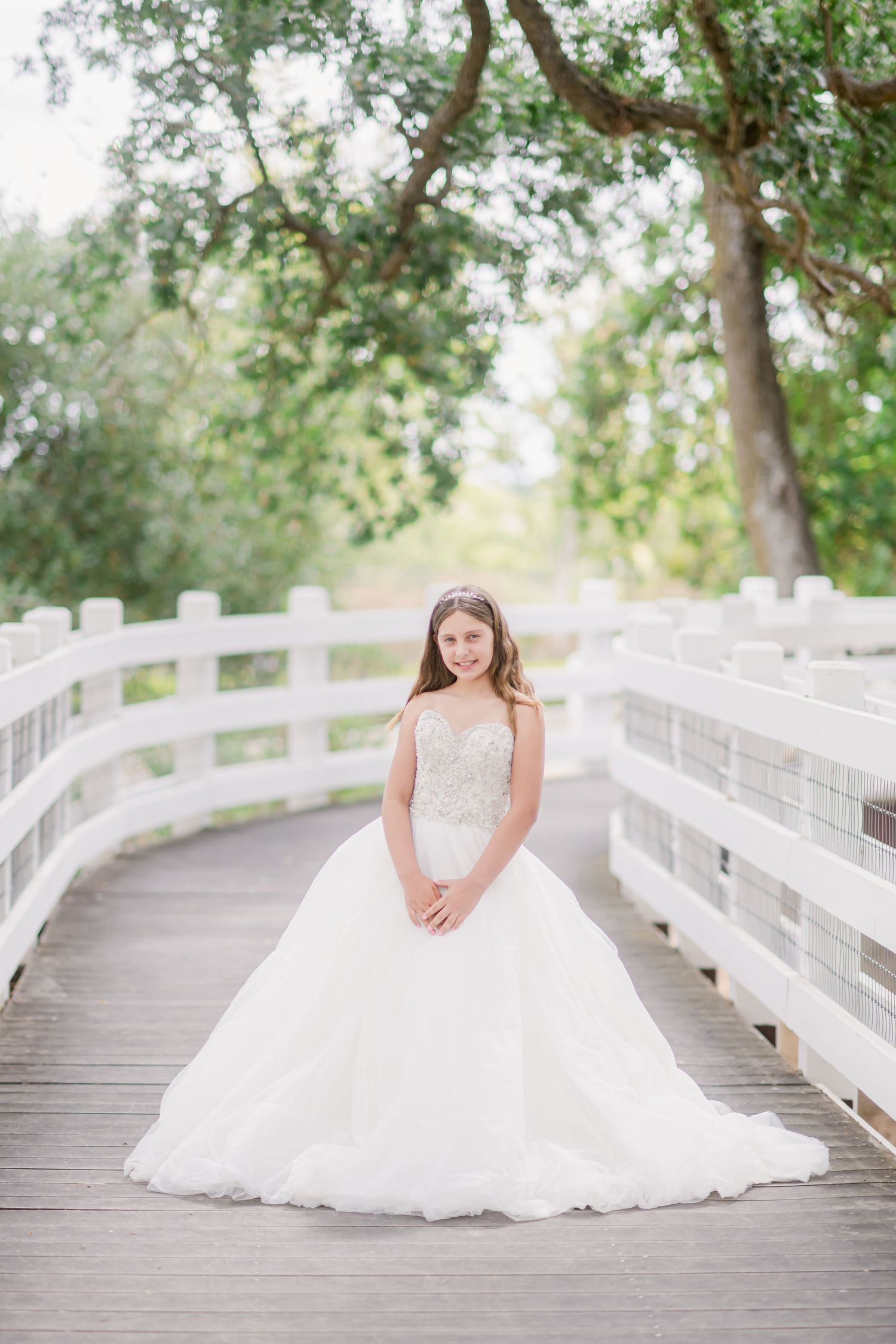 A bride in a flowing white wedding gown sits gracefully on a white wooden bridge surrounded by lush greenery.