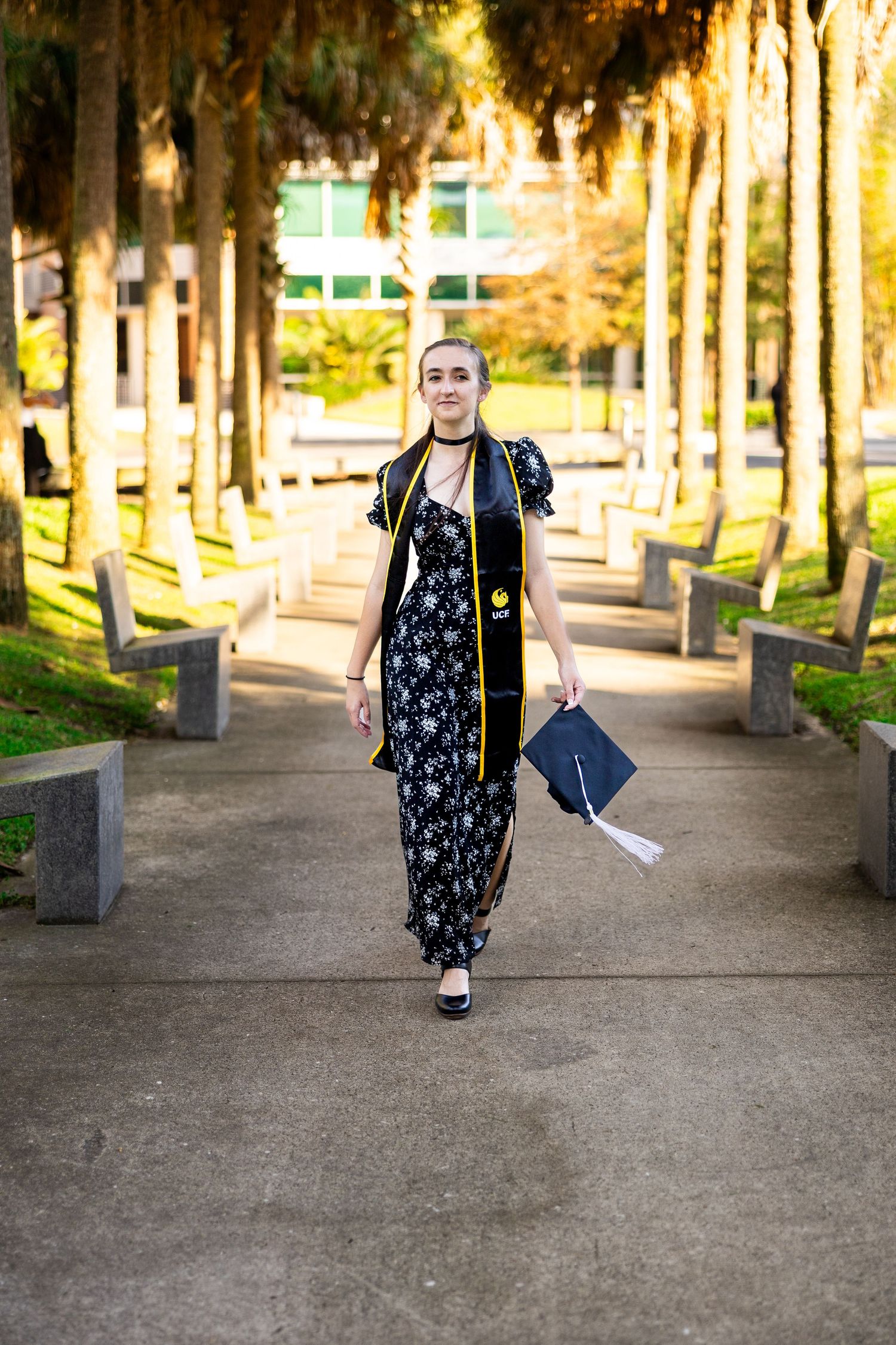 Graduate walking down tree-lined path in black cap and gown holding diploma.