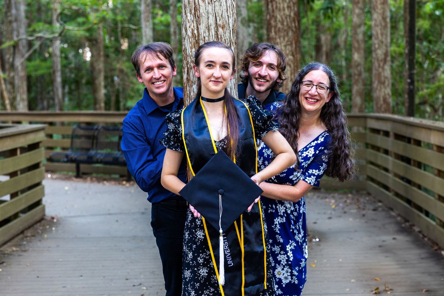 Graduate celebrates with friends on wooden boardwalk surrounded by forest.