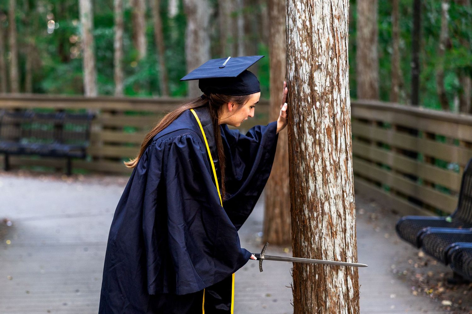 Graduate in navy blue cap and gown walking among tall trees.