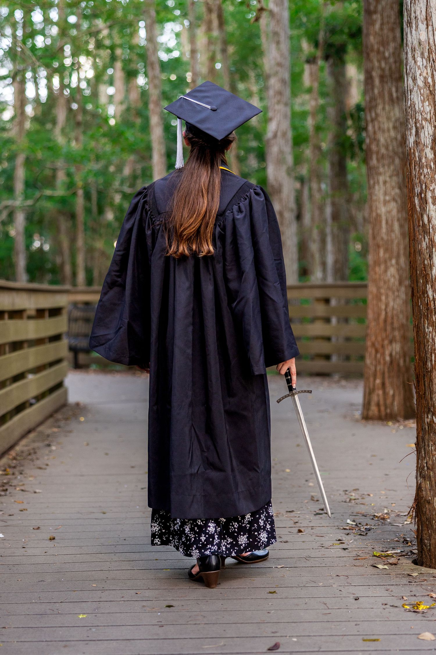 Graduate in black cap and gown stands on wooden boardwalk path through wooded area.