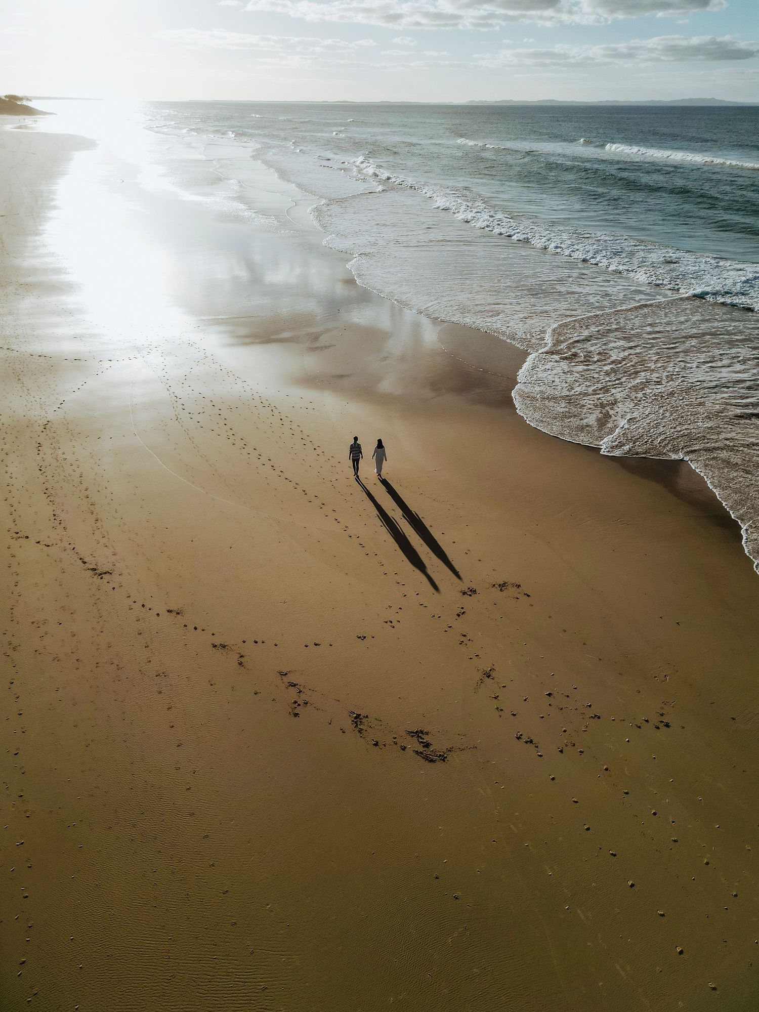 Two silhouetted figures cast long shadows while walking along a sandy beach at sunset with gentle waves rolling in.