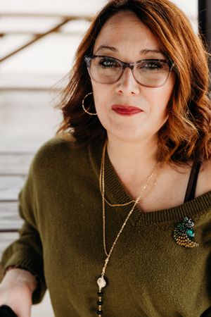 Portrait in olive green sweater wearing cat-eye glasses, red lipstick, and layered necklaces with decorative brooch.