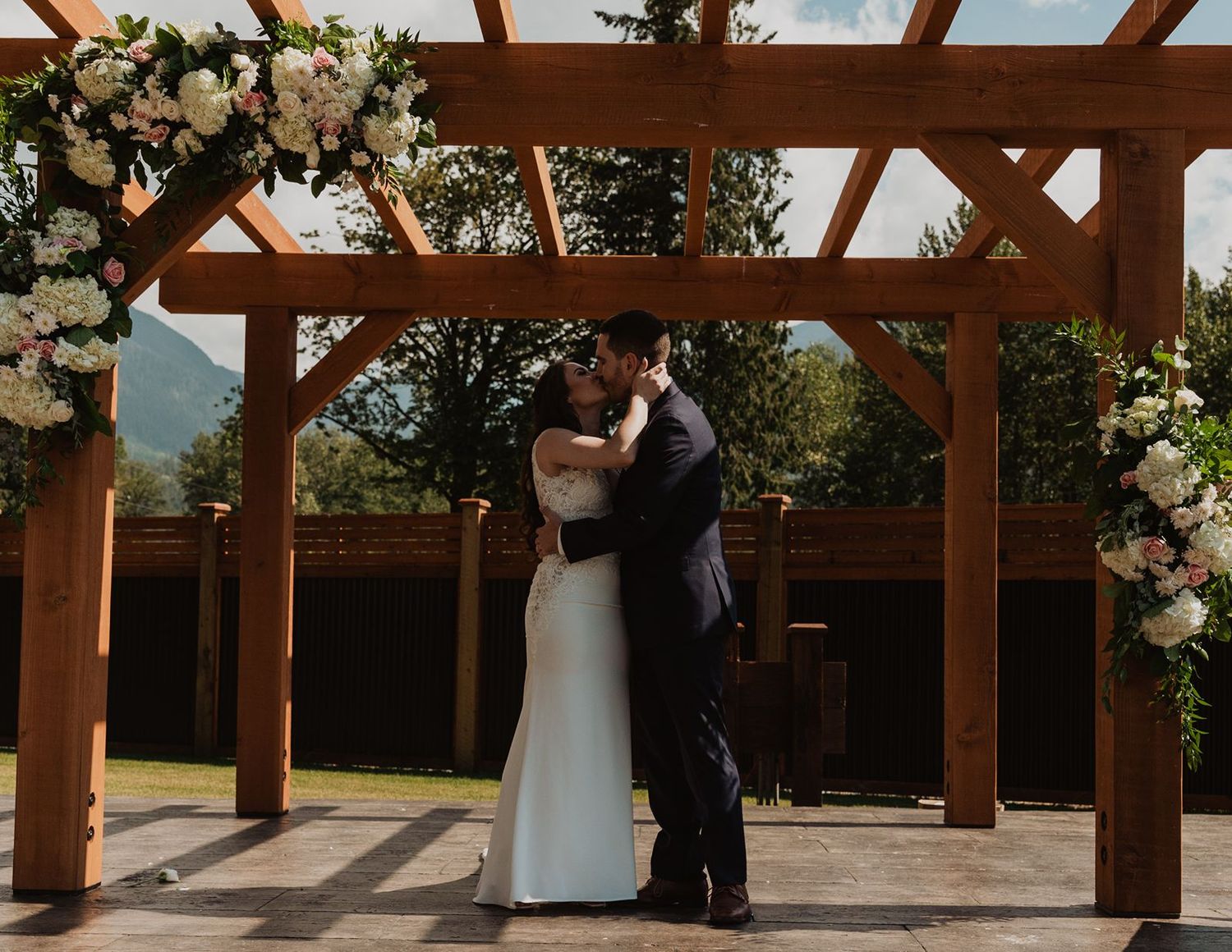 Bride and groom sharing first kiss under a wooden pergola adorned with white and pink flowers.
