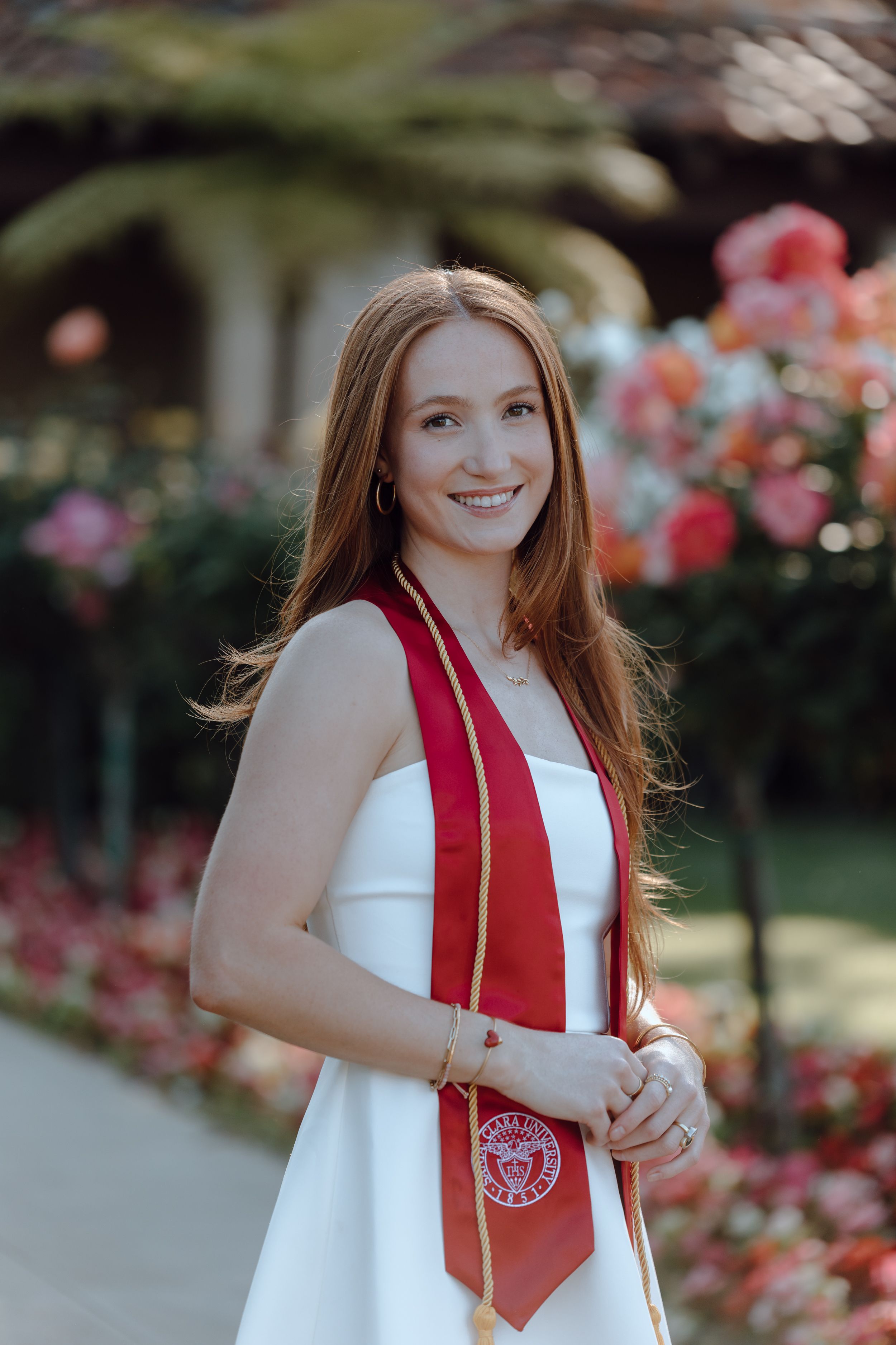 Graduate in white dress with red stole poses for portraits in a garden with blooming flowers in the background.