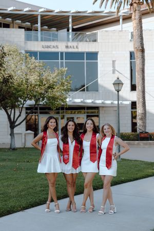 Four college graduates stand and pose together for their grad photoshoot at Santa Clara University. Outside their major's building on campus