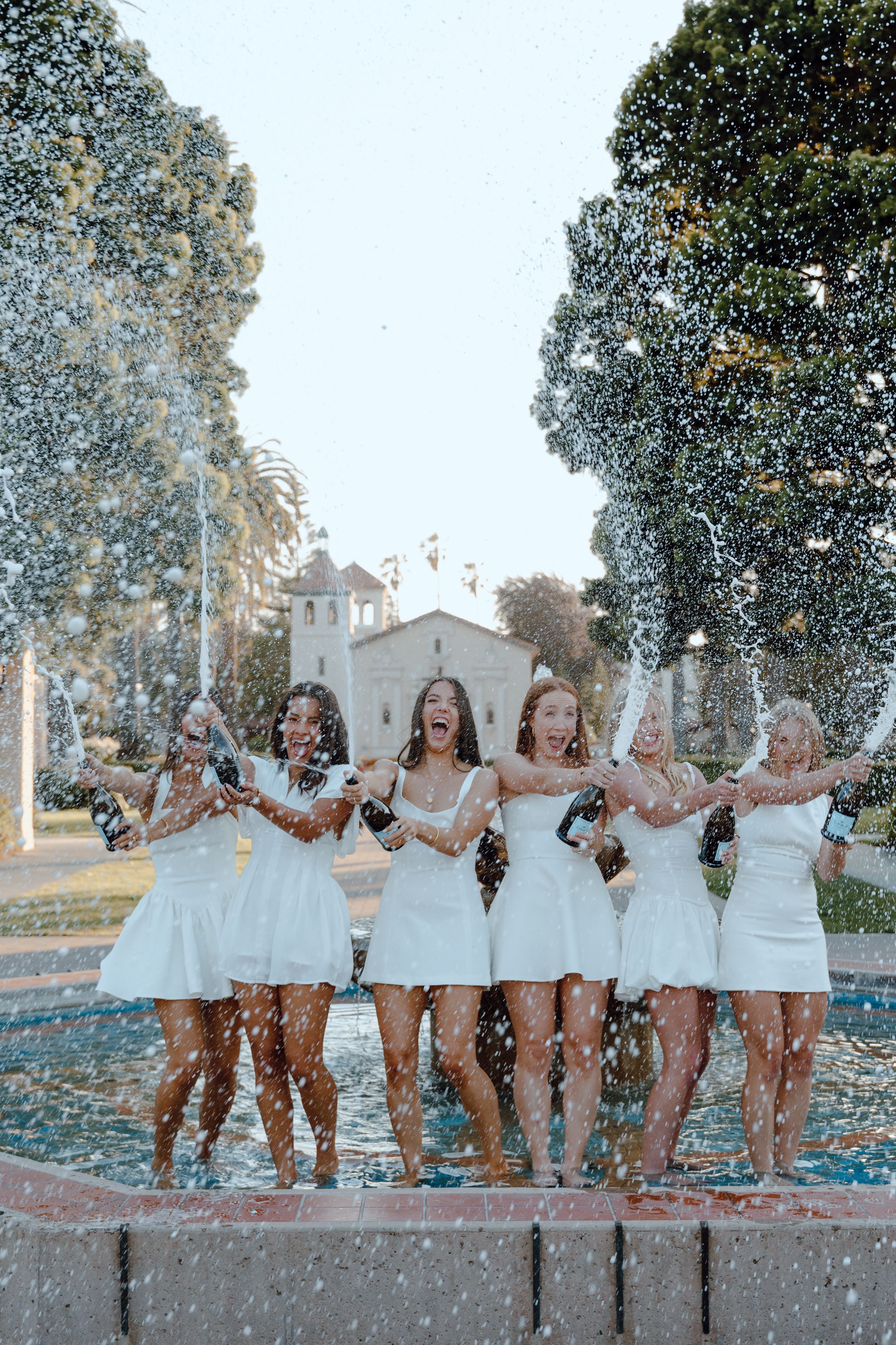 Group of college graduates pop and spray champagne while they stand in a fountain for their grad photoshoot at Santa Clara University