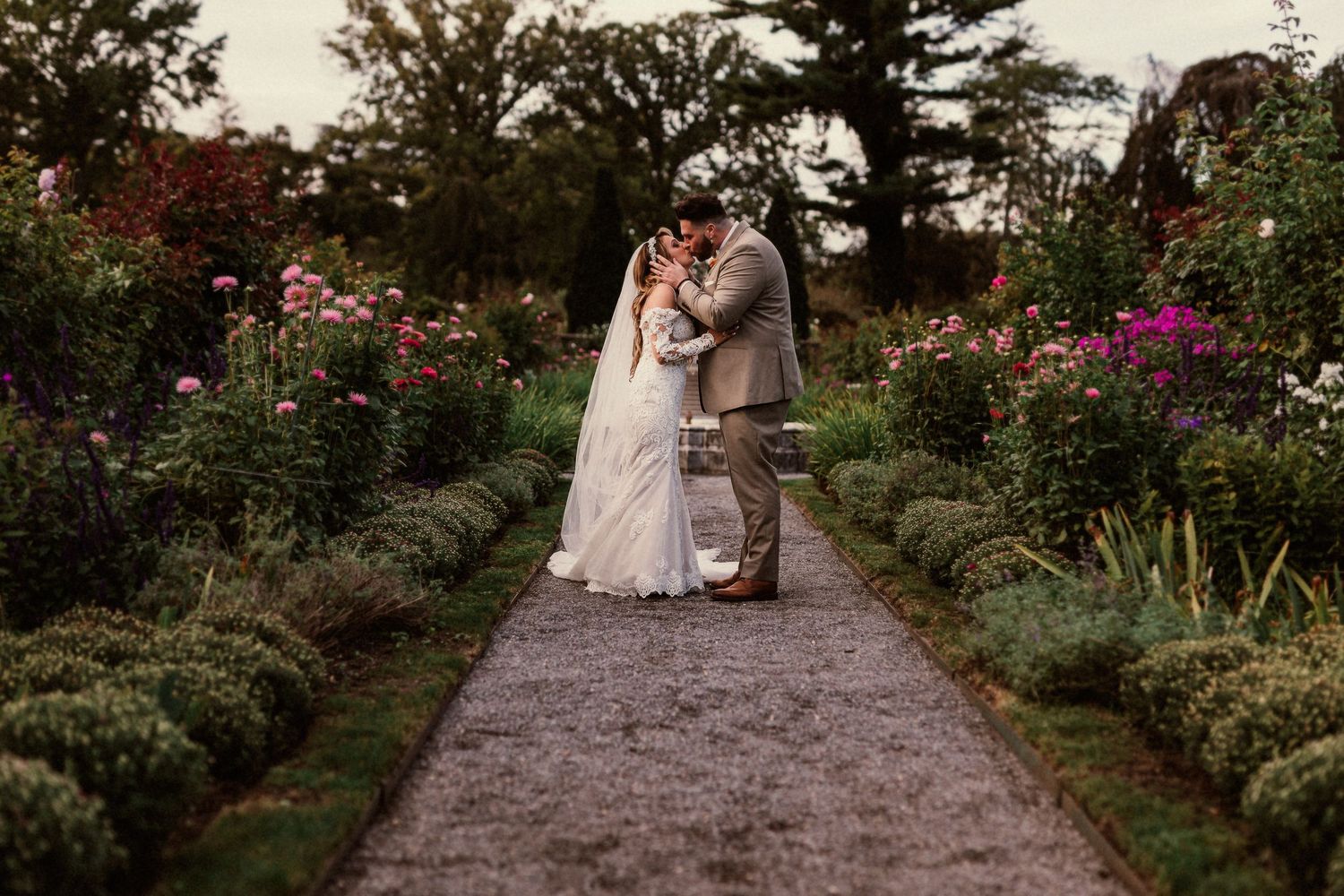 A wedding couple poses on a garden path surrounded by vibrant flowers and greenery.