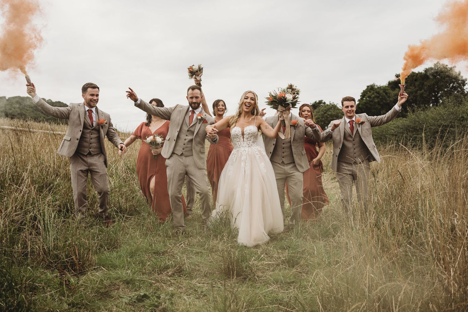 Bride and groom celebrating with their wedding party in a field, walking together holding smoke flares