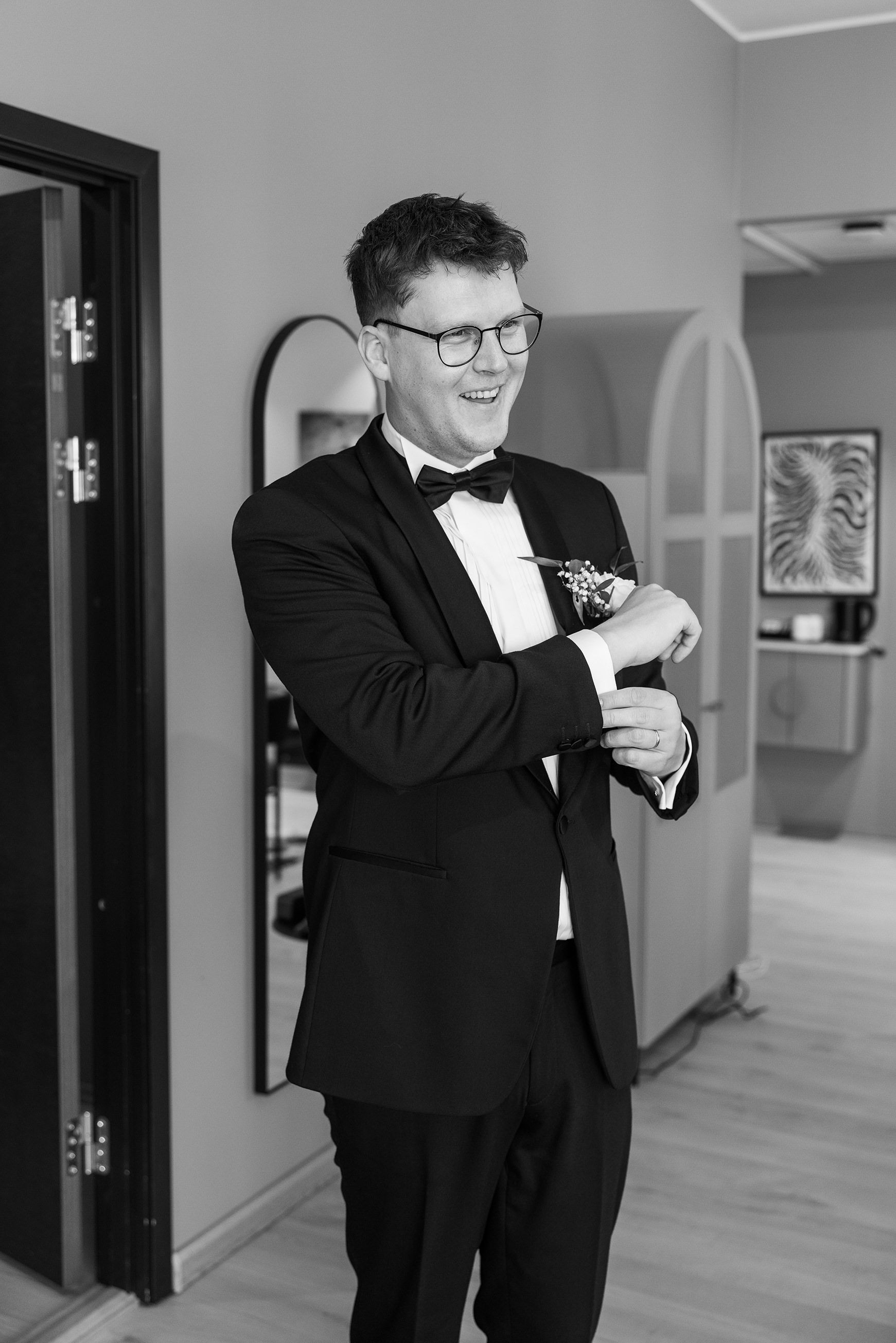 Sort-hvitt bilde av brudgom som retter litt på skjorta på et hotellrom.