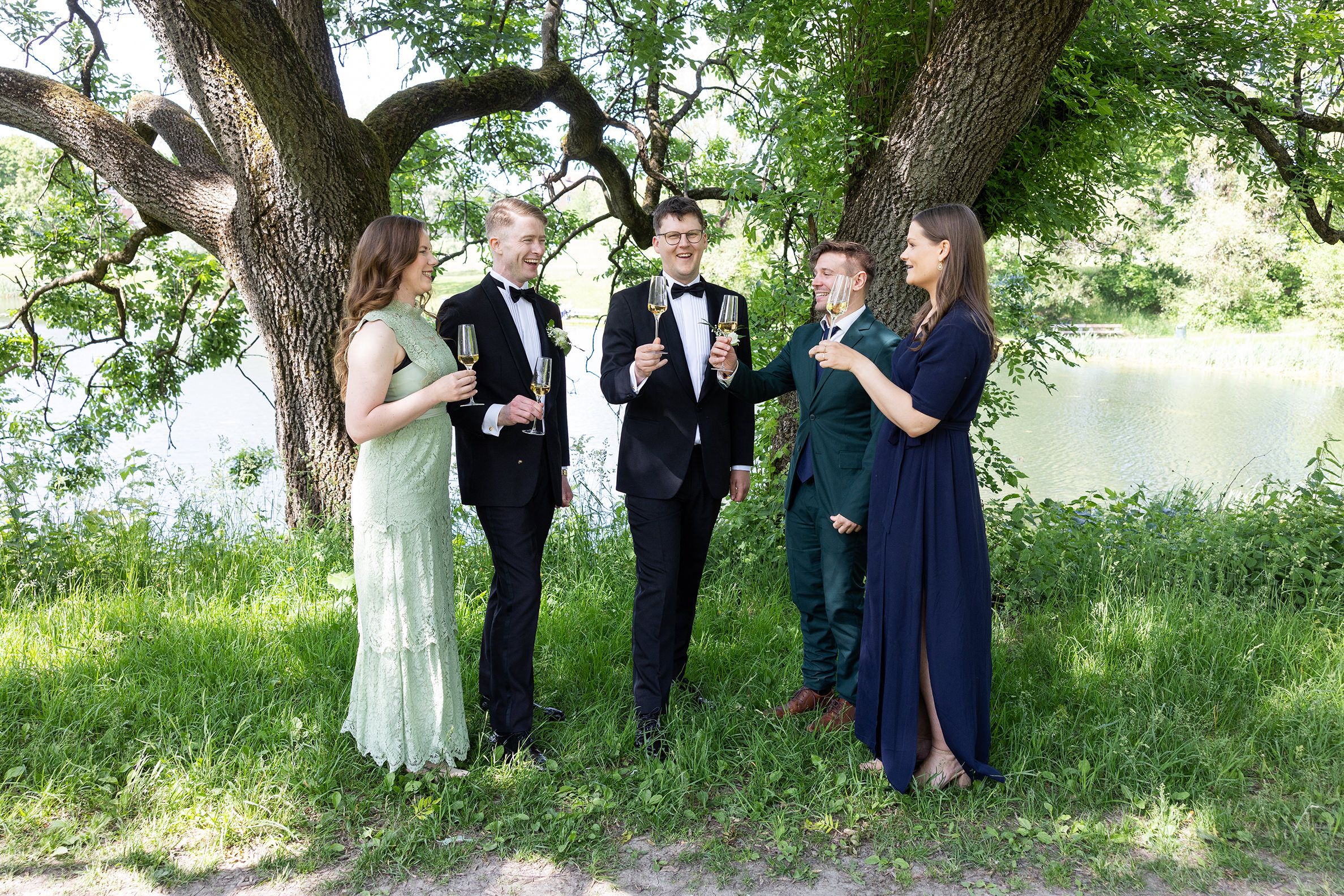 Forlovere og burdepar stående i en park med grønne trær. De prater, holder et glass champagne og skåler.