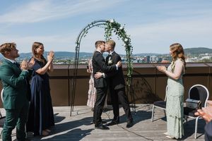 Brudepar som kysser foran en blomsterbue og utsikt over Oslo. Forlovere står rundt og klapper.