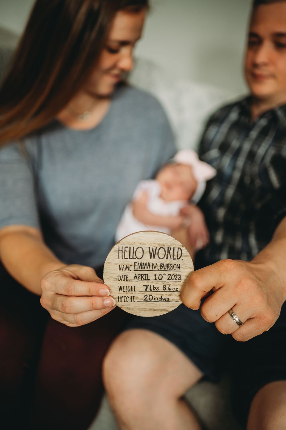 A couple holds up a wooden birth announcement sign reading 'Hello World' while sitting with their newborn baby.