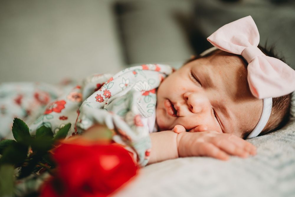 A sleeping newborn baby wearing floral clothing rests peacefully with a red flower accent in the frame.