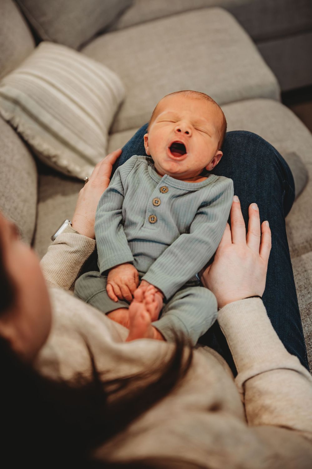A sleeping newborn baby wearing a gray onesie yawns while resting on soft bedding.
