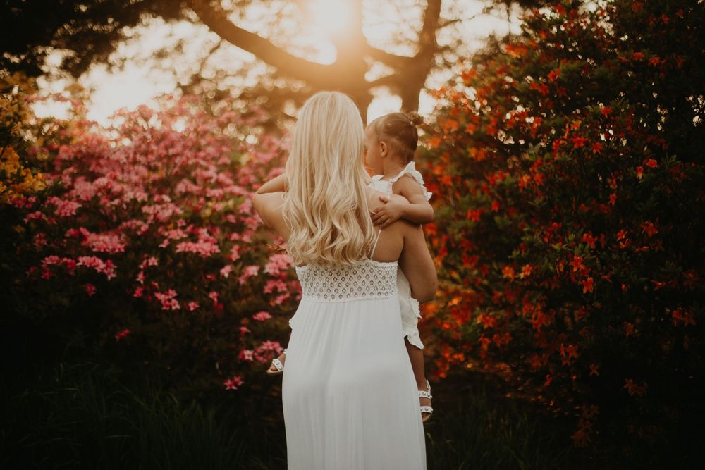 A white-dressed figure holds a baby against a backdrop of vibrant pink azalea bushes at golden hour.