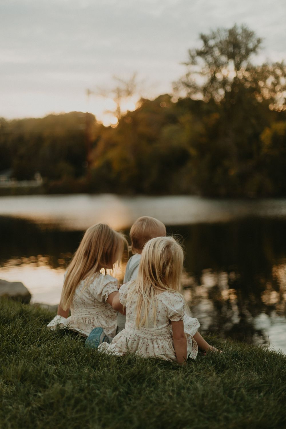 Three children in white dresses sit by a lake at sunset enjoying the peaceful view.