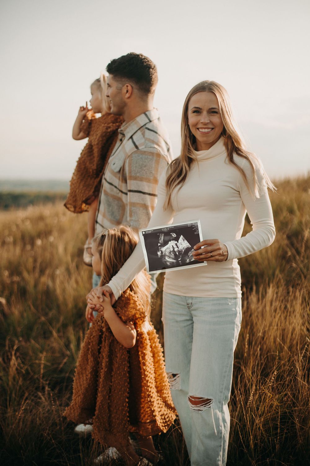 A family stands in a golden field at sunset while holding an ultrasound photo, suggesting a pregnancy announcement.