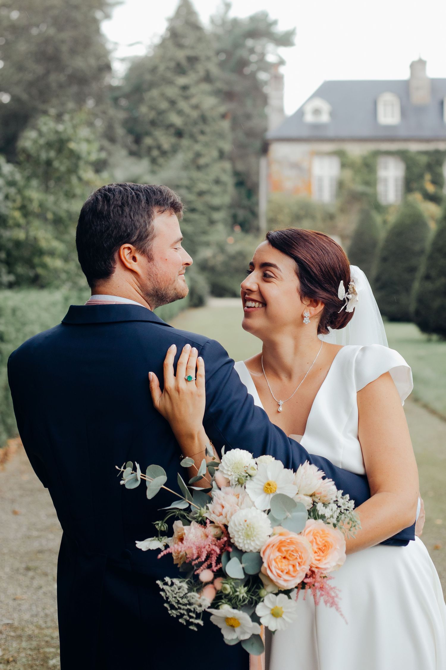 Smiling bride holding pastel bouquet gazes at groom in navy suit in lush garden setting.