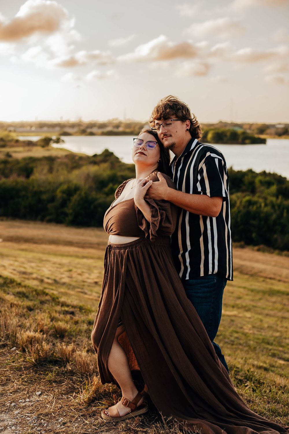 A couple embraces tenderly in a field at sunset, with rolling hills and trees in the background.