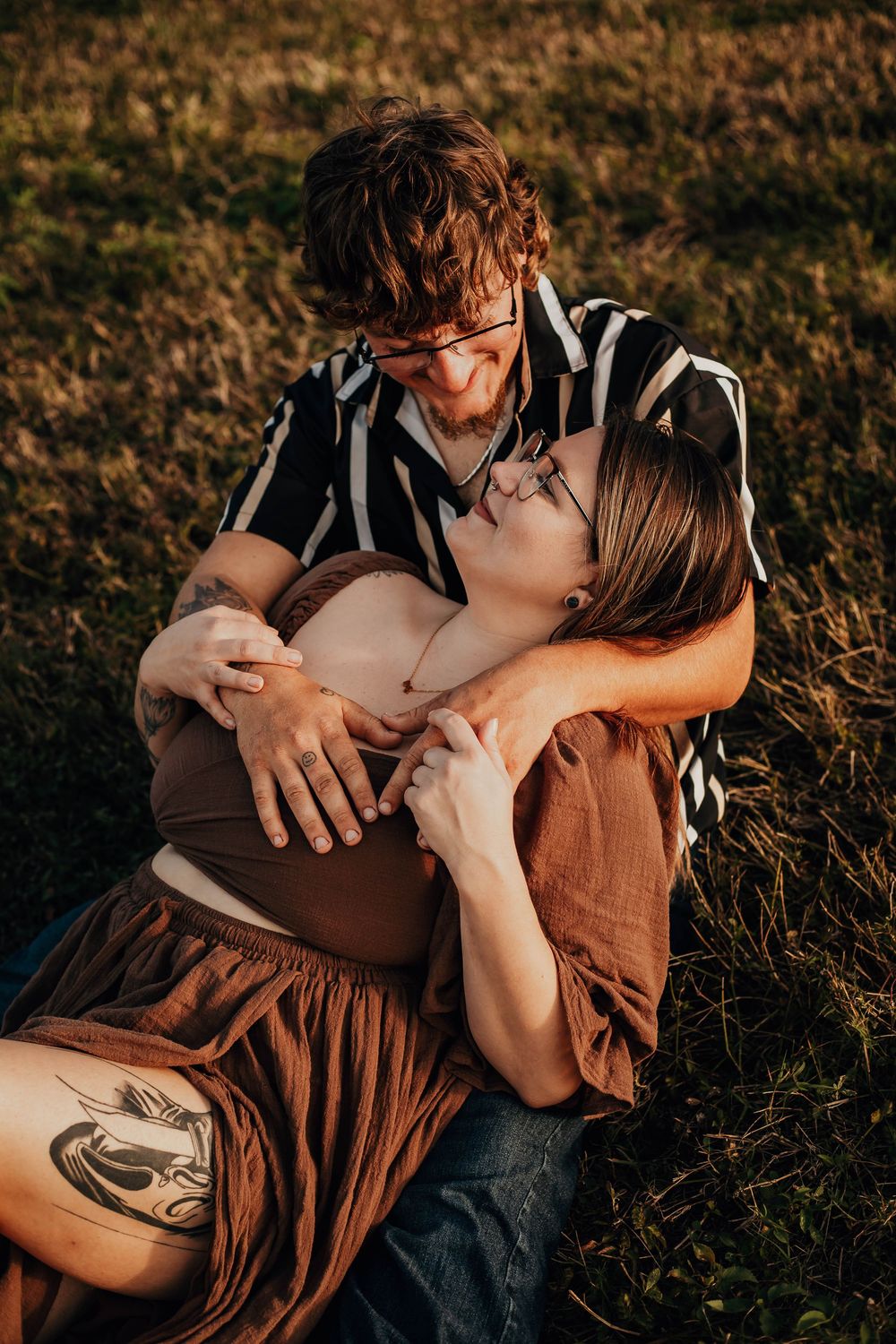 Romantic couple in casual attire cuddles together on grass during sunset photoshoot.