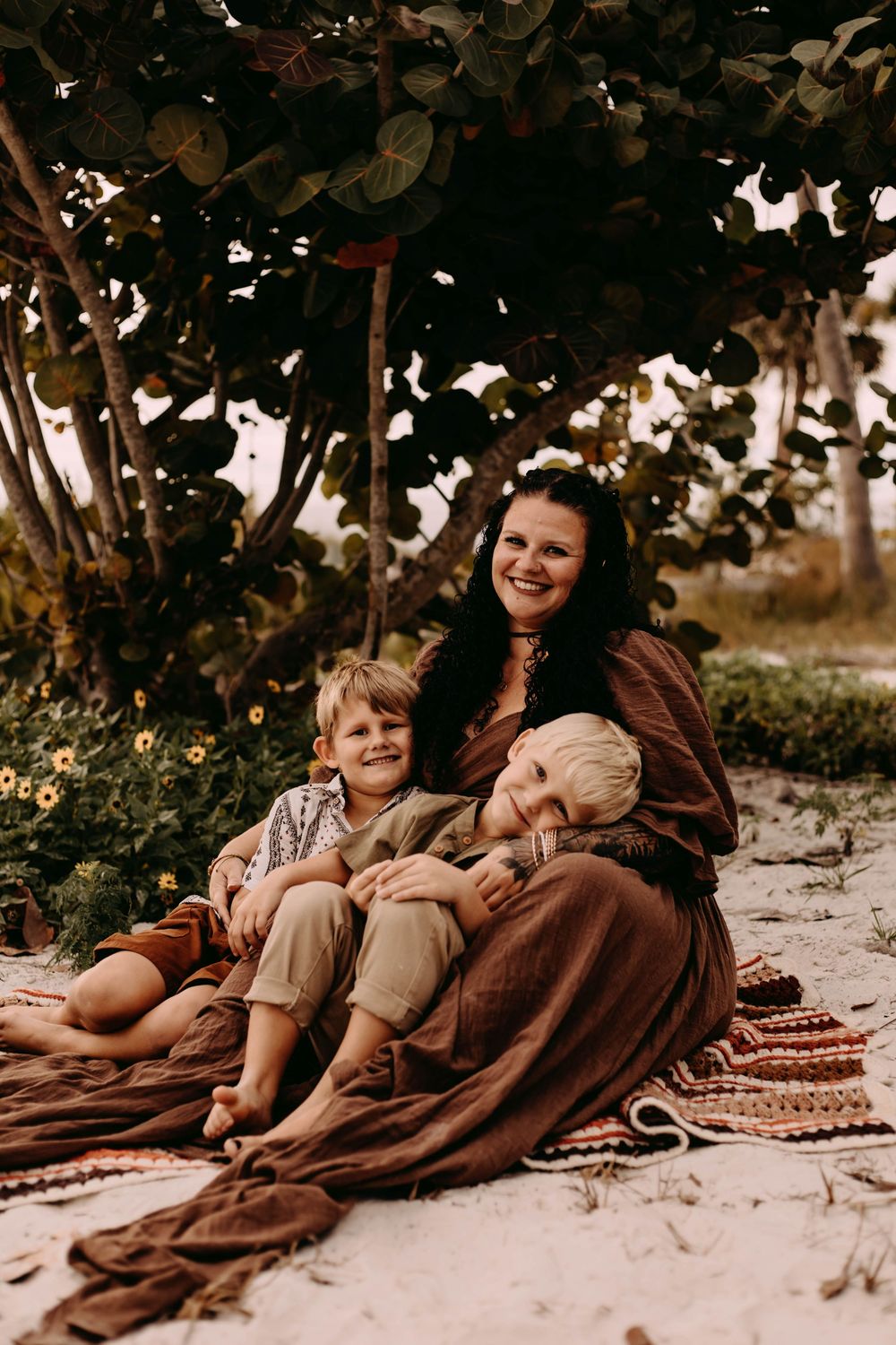 A family relaxes together on a brown blanket under trees during an outdoor photo session at sunset.