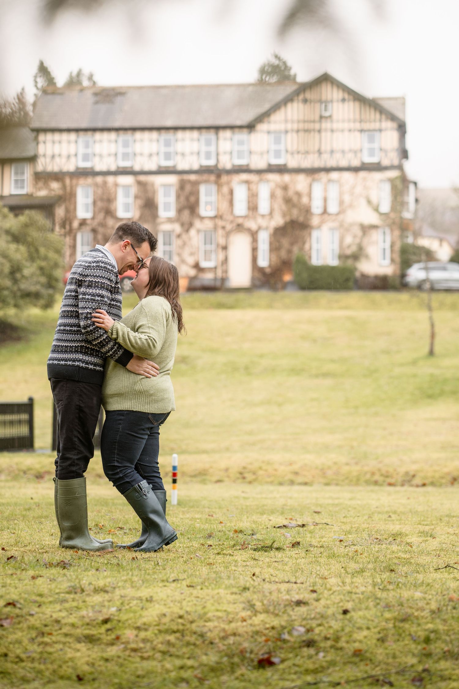 A couple shares a tender moment on a grassy hill with stone buildings in the background on an overcast day.