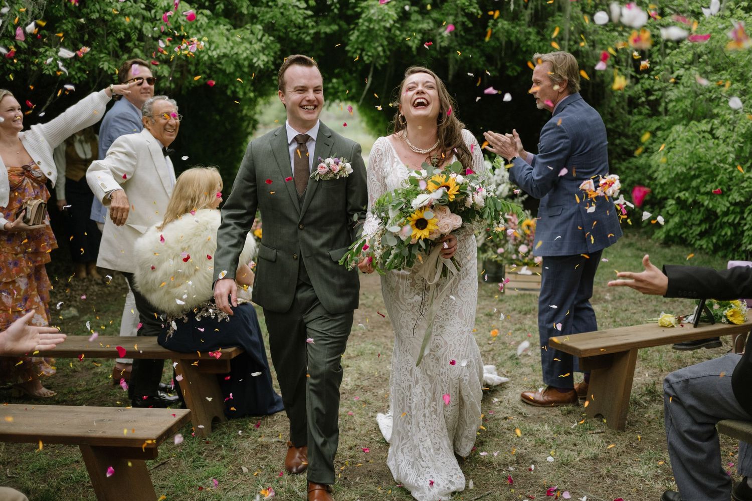 A joyful celebration in a garden setting with wedding flowers and guests gathered around wooden benches.
