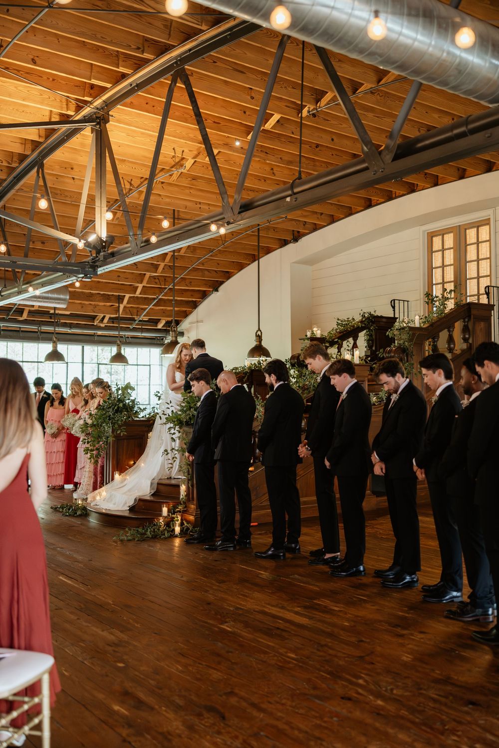 Wedding guests line the sides of a spacious industrial venue with exposed wooden beams and string lights overhead at Summerour Studio