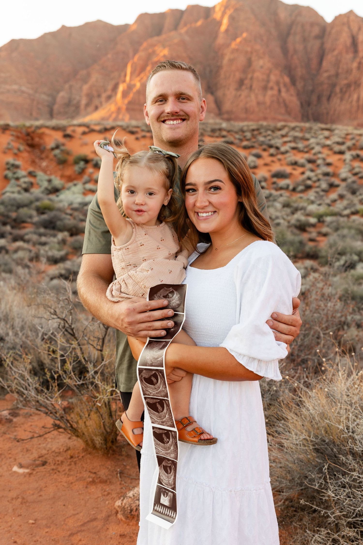 St. George Utah family photographer capturing mom, dad, and 2-year-old daughter smiling together while holding a sonogram in desert landscap
