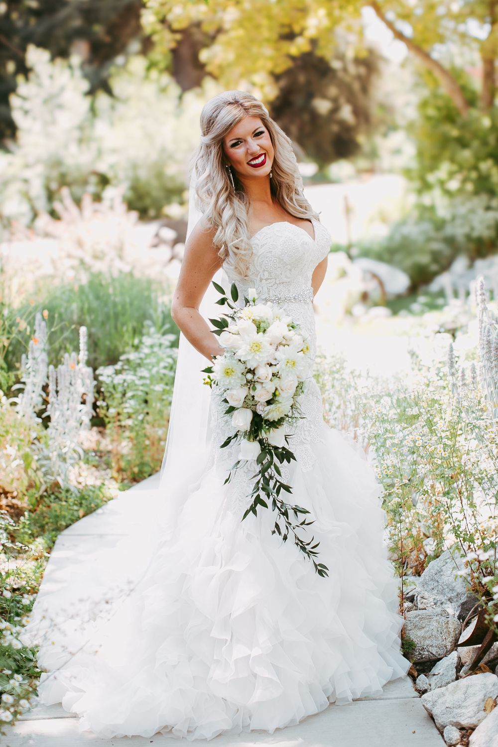 A bride in a flowing white ballgown poses joyfully in a garden setting with delicate white flowers and greenery.