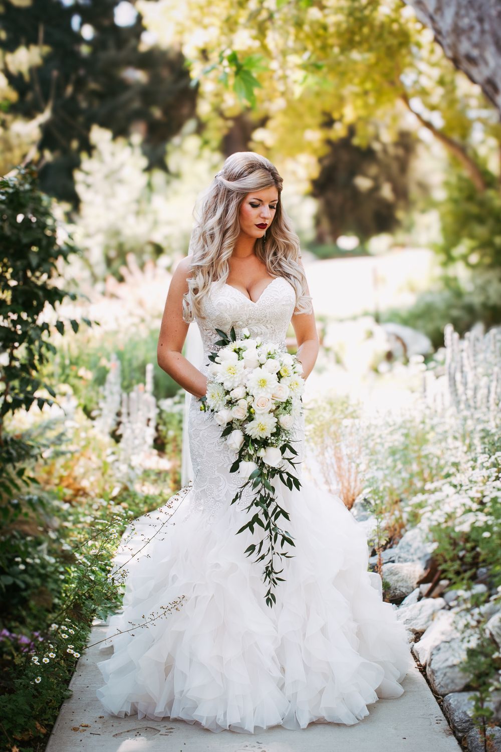 A bride in a flowing white gown poses with a cascading white bouquet in a sunlit garden setting.