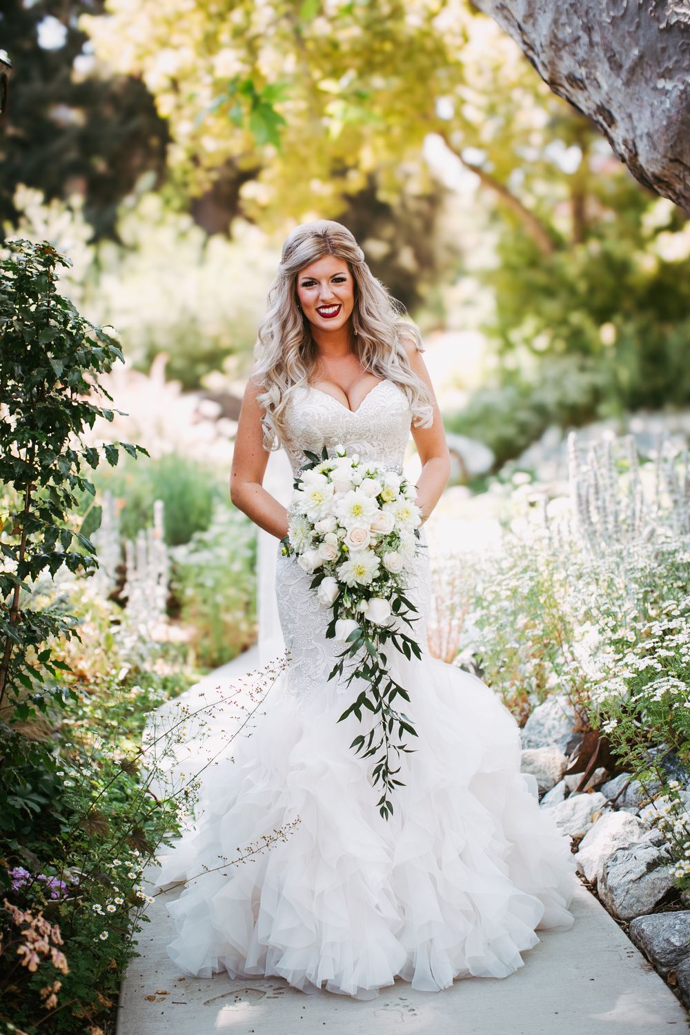 A bride in a flowing white wedding gown poses in a garden setting surrounded by delicate white flowers and greenery.