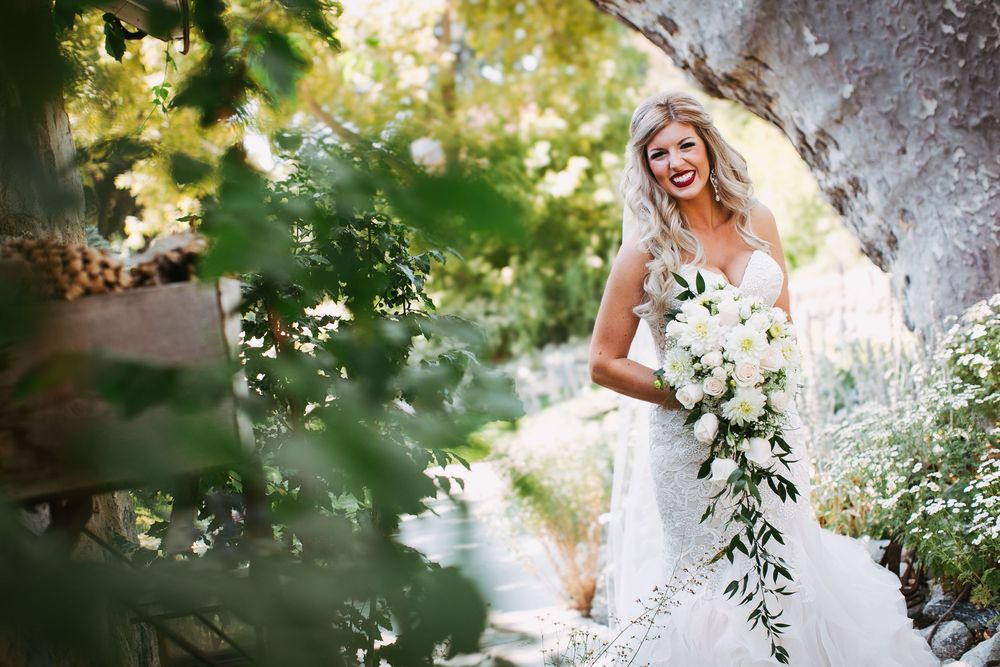 A bride in a white lace gown holds a cascading bouquet of white flowers in a dreamy garden setting.