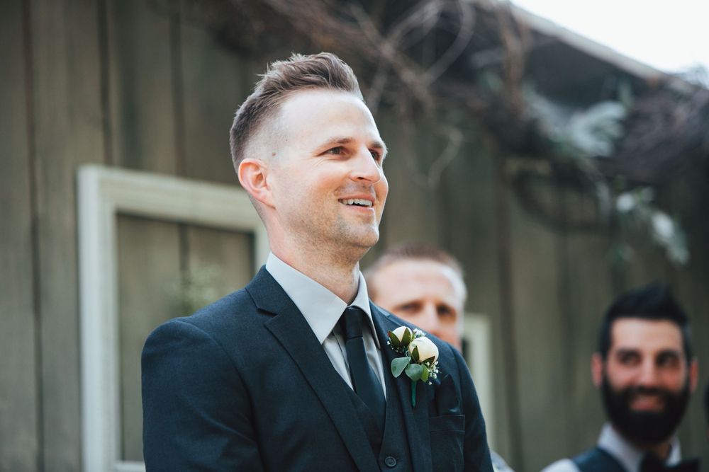 A joyful person in a black suit and tie with a floral boutonniere smiles during an outdoor wedding ceremony.