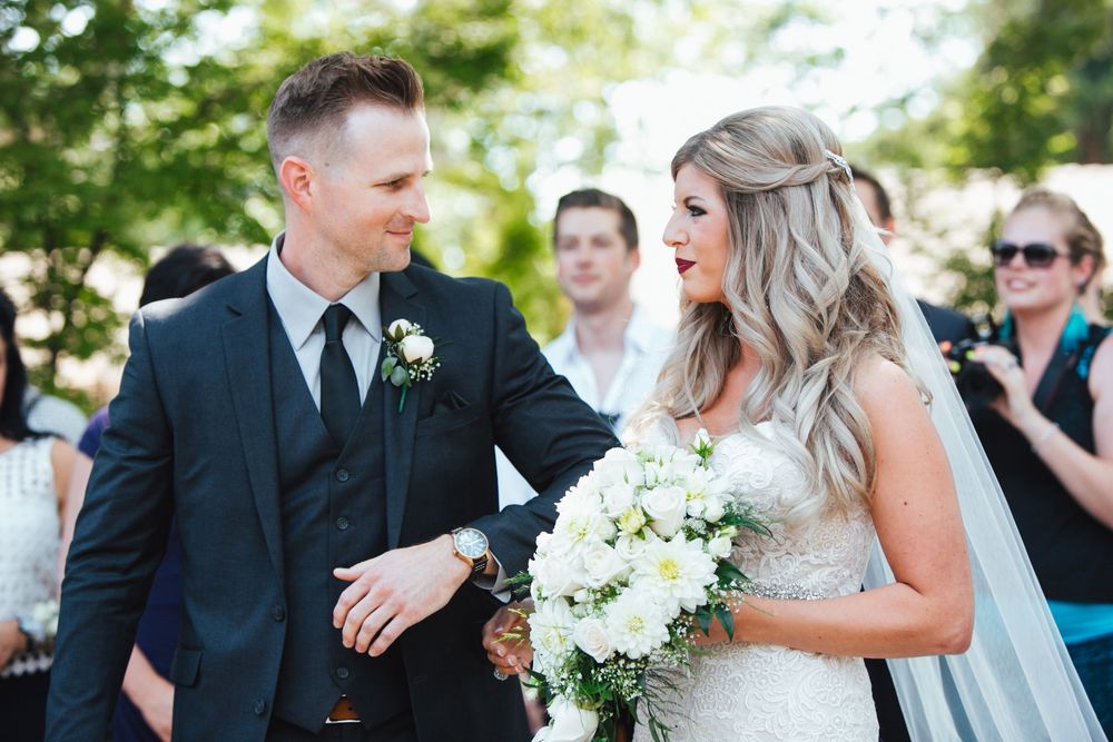 Wedding party celebrates outdoors on sunny day with formal attire and white floral bouquets.