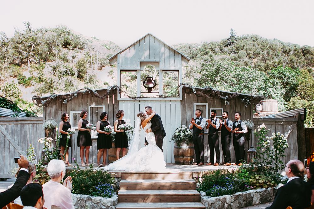A rustic barn wedding ceremony setup with wooden benches and floral arrangements in a garden setting.
