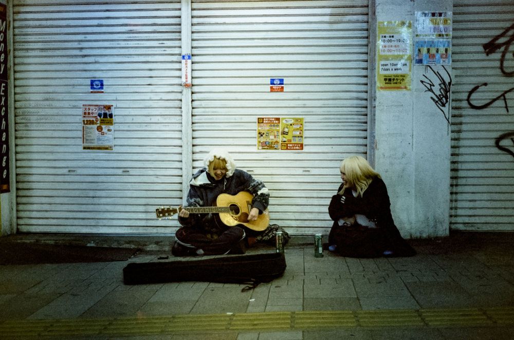 Two street musicians perform with a guitar case open for tips in front of closed metal shutters at night.
