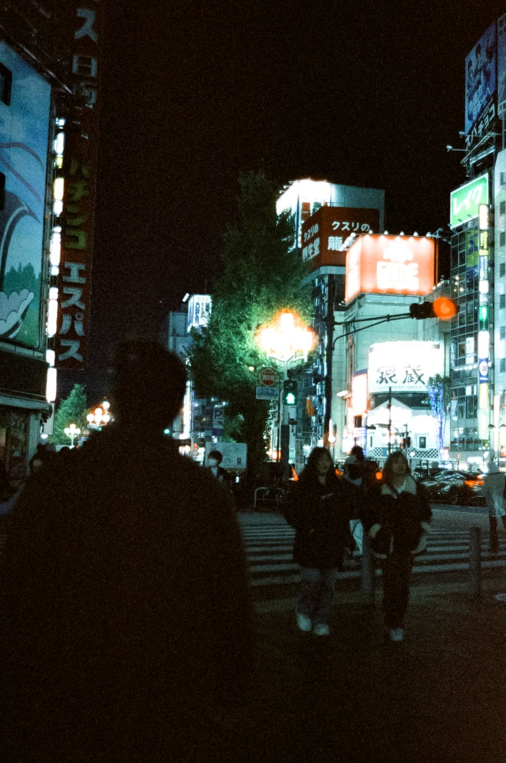 Dark urban street scene at night with illuminated storefronts and silhouettes of people walking on the sidewalk.