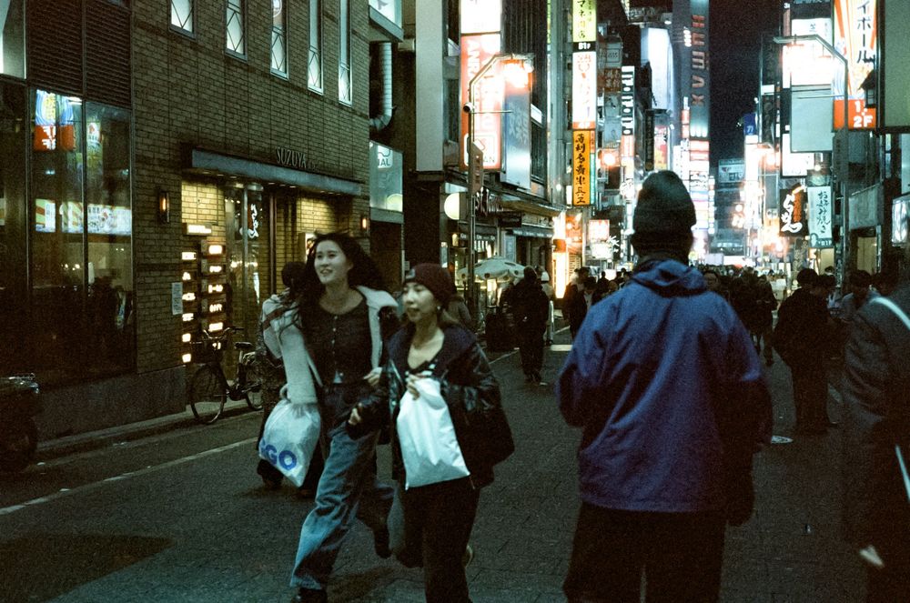 People walking on a city street at dusk with illuminated storefronts and neon signs creating an urban atmosphere.