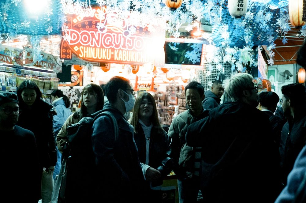A crowded night market scene with neon signs and people gathered around a donut stand.