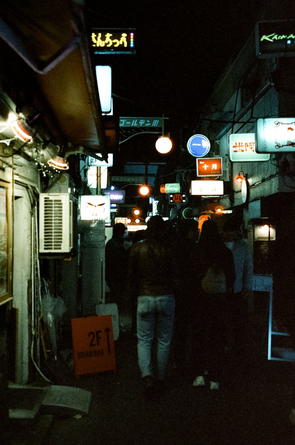 Dimly lit narrow alley in Japan with neon signs and air conditioning units mounted on dark building walls.
