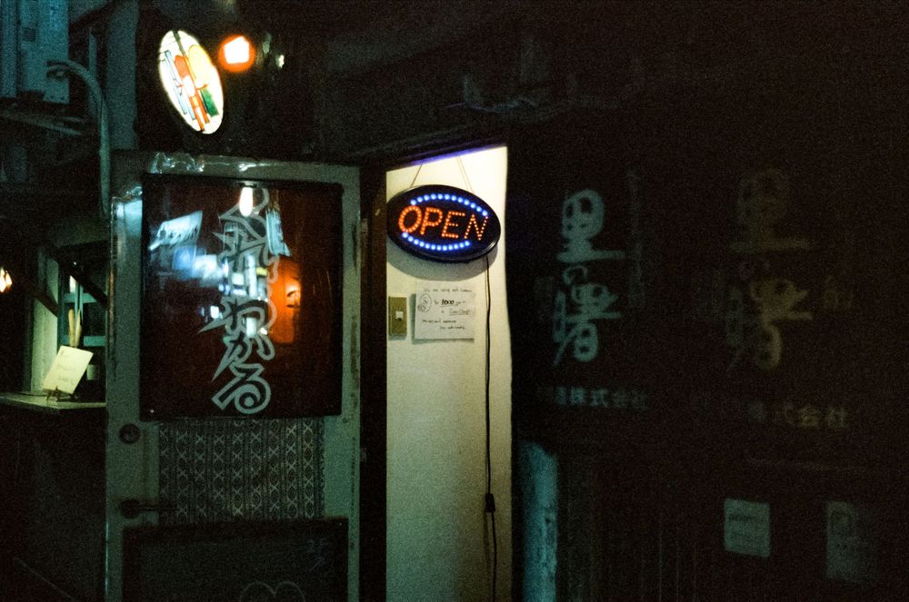 Dark storefront with neon signs and illuminated windows casting a moody glow on a nighttime street.