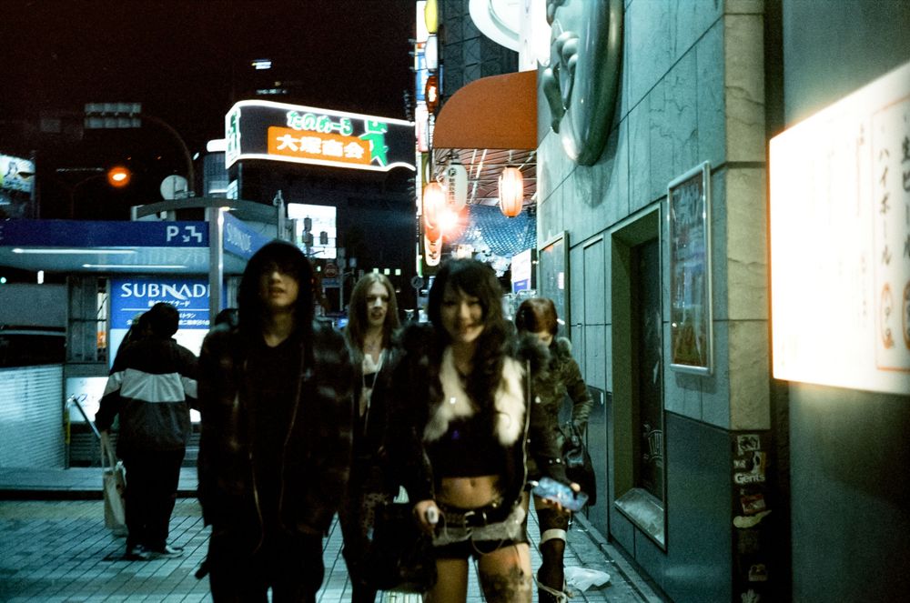 Cinematic nighttime street scene in Japan with neon signs and people walking past illuminated storefronts.