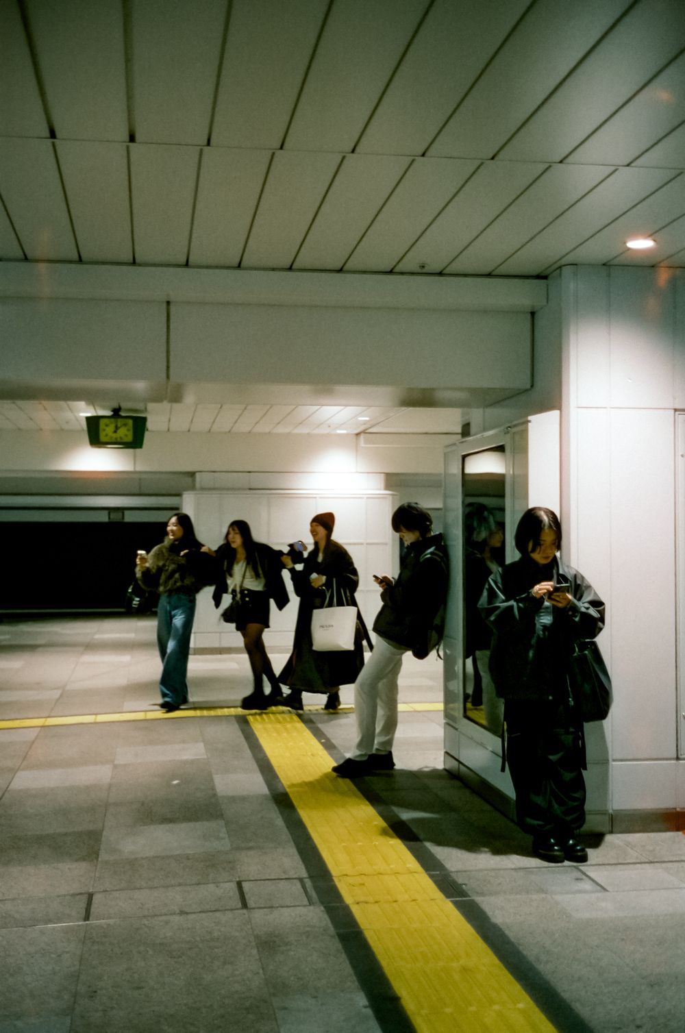 People waiting along a yellow guideline in a modern subway station corridor with fluorescent lighting overhead.