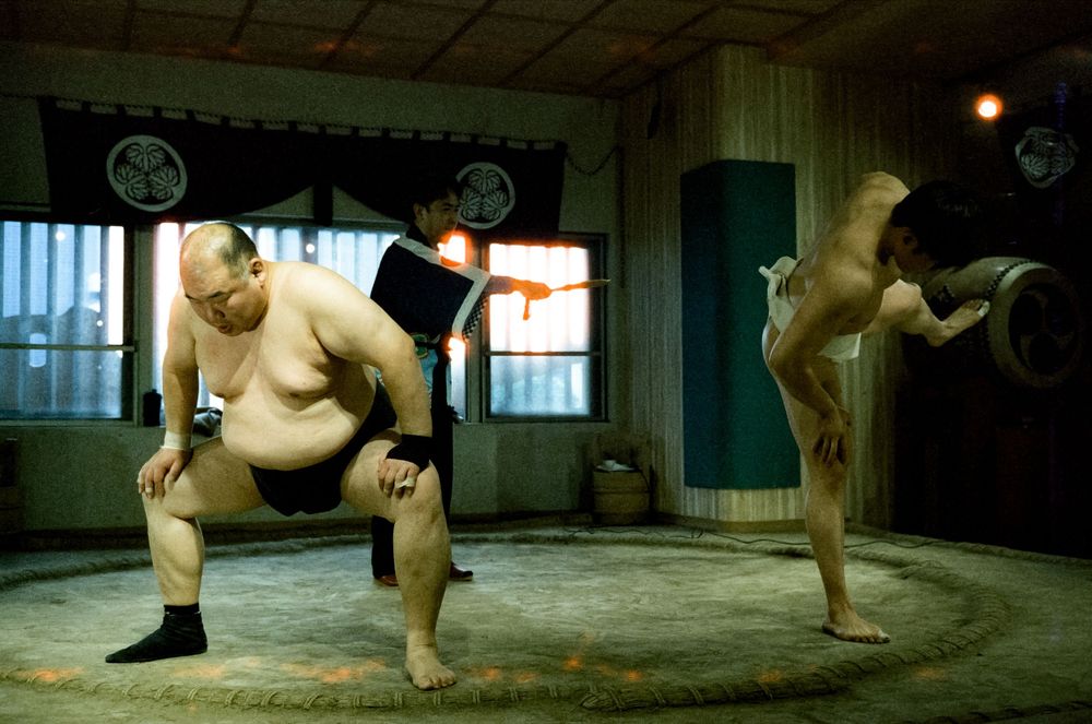 Two sumo wrestlers training in a traditional dojo with one performing a high kick while the other stands in stance position.