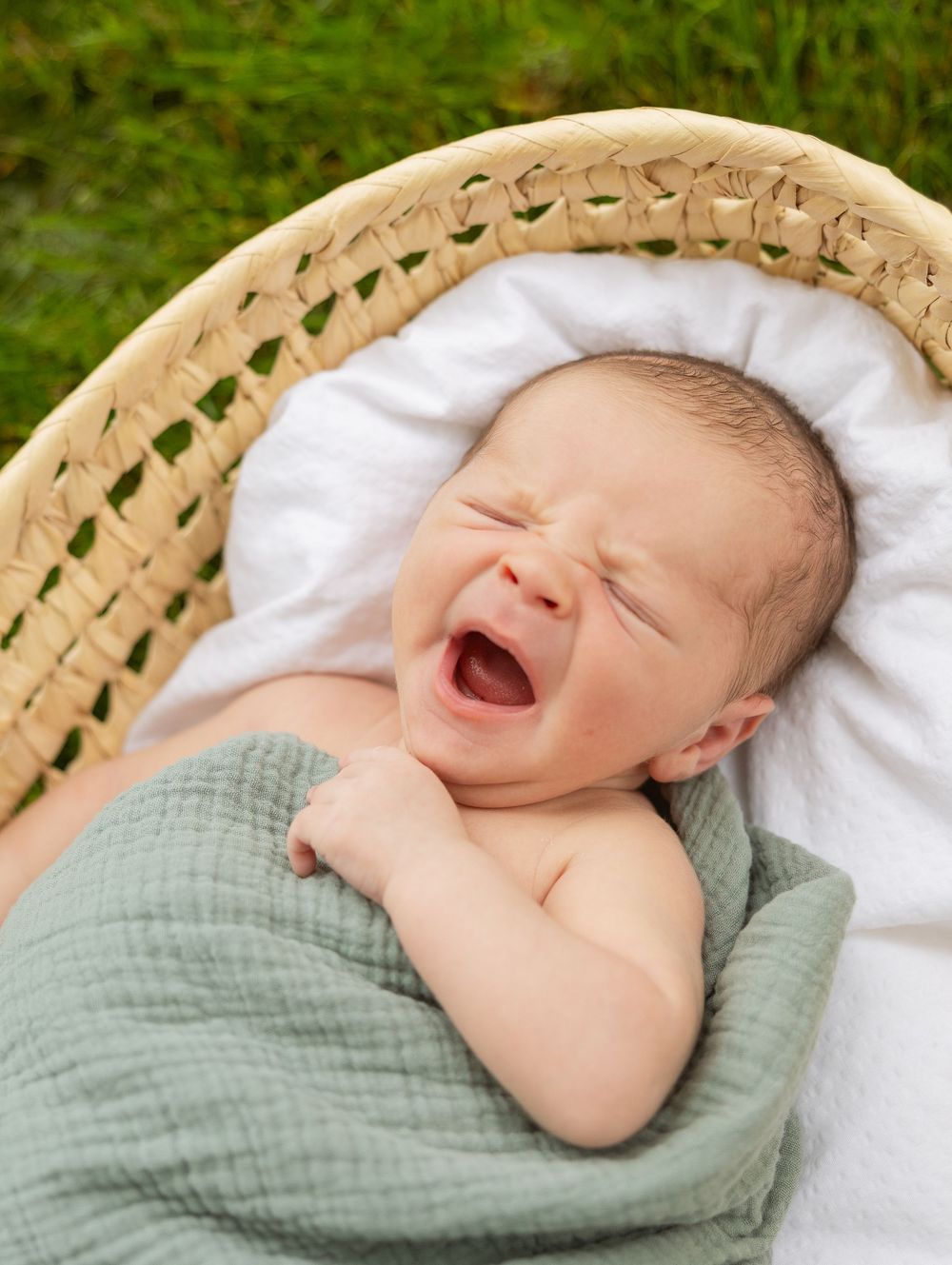 Newborn baby yawning while wrapped in sage green muslin blanket in woven basket.